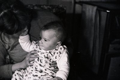 A black and white photo captures a baby held by an adult. The child wears a patterned onesie, gazing directly at the camera. ...