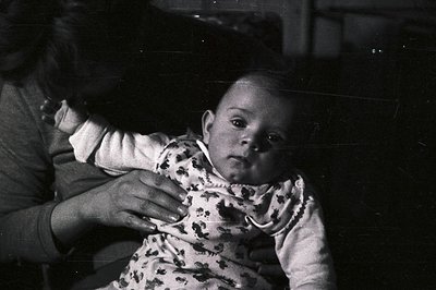 Black and white portrait of a baby held by an adult. The child wears a patterned romper, looking directly at the camera. A ha...