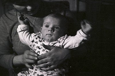 A grayscale image depicts a baby held by an unseen person. The infant, wearing a patterned romper, gazes directly at the came...