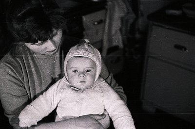 A mother cradles an infant bundled in a soft, textured bonnet and sweater. Interior scene with a dresser and partially visibl...