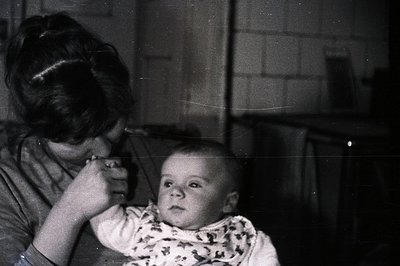 A mother tenderly feeds a baby in a dimly lit interior, likely a kitchen. The baby wears a patterned bib, looking directly at...