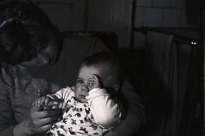 A black and white image captures a young child held by an adult, likely a parent. The child, dressed in a patterned onesie, r...