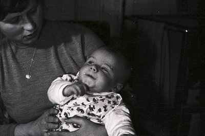 A close-up, black & white photograph captures a baby held by an adult. The baby, dressed in a patterned romper, gazes upwards...