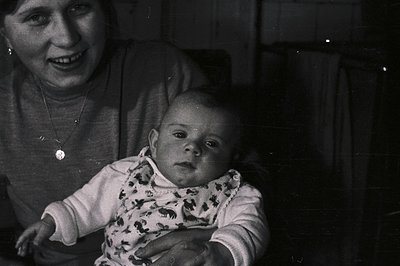 A young mother smiles gently as she holds a baby with wide, observant eyes. The baby wears a patterned bib. Likely a candid f...