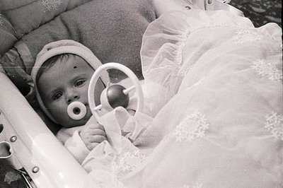 Infant in a vintage carriage, pacifier visible, surrounded by ornate lace and cushioned padding. Likely 1950s-1970s, suggesti...