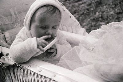 Infant seated in a wicker basket, wearing a bonnet and long-sleeved dress. Appears to be chewing on a teether. Likely a candi...