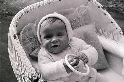 Infant in a wicker bassinet, adorned with a bonnet and knit romper suit. Small hands grip a toy steering wheel. Likely a fami...