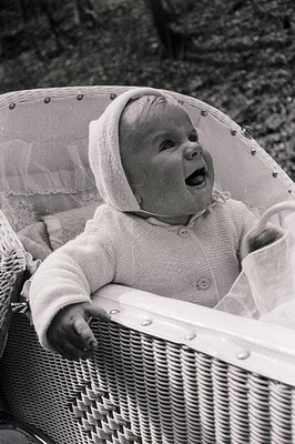 A baby, bundled in a knitted bonnet and sweater, expresses joyful surprise from within a wicker baby carriage. Classic mid-ce...