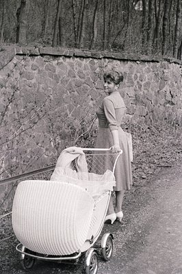 A woman in a mid-length dress and heels stands beside a vintage pram with elaborate metalwork and fabric canopy. The setting ...