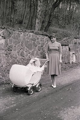 A woman walks along a paved path, pushing an elaborate wicker baby carriage. She wears a modest, knee-length dress and appear...