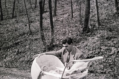 A man in a suit pushes a wicker baby carriage through a wooded area with fallen leaves. The infant within is sleeping. Likely...