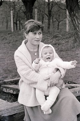 A woman in a crocheted cardigan and dress holds a baby wearing a matching outfit and bonnet. They sit on a stone bench in an ...