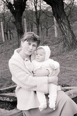 A woman holds an infant, both wearing matching knitted suits with hoods. Seated on a wooden structure near a gently sloping, ...