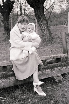 A woman in a knee-length skirt and heels cradles a baby bundled in a knit outfit. Seated on a rustic wooden barrier, they are...