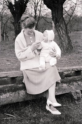 A woman sits on a wooden fence, holding a baby bundled in white. The woman wears a fitted dress suit and pointed heels, indic...