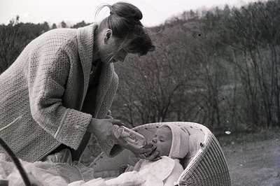 A woman in a cardigan gently feeds a baby in a wicker basket outdoors. The setting appears to be a garden or park with a blur...