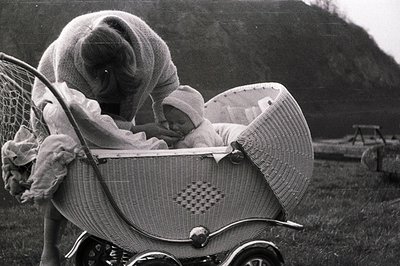 A young baby rests in an elaborate wicker pram, likely mid-century. The mother, bundled in a large sweater, leans over the in...