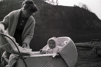 A young mother stands near a wicker baby carriage containing an infant in a bonnet, posed outdoors on a slight incline. The s...