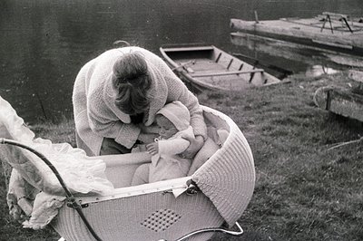 A woman in a crocheted shawl tends to an infant nestled in an ornate, woven wicker crib beside a body of water. The scene evo...