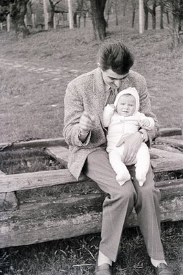 A man in a tweed jacket and tie cradles an infant dressed in white. Seated on a weathered wooden fence, they appear outdoors ...