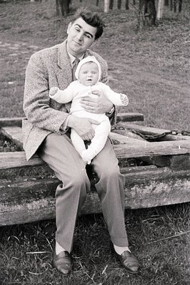 Man in a tweed jacket and trousers cradles a baby wrapped in white, seated on a wooden structure, possibly a bench. Appears t...