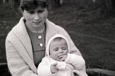 A young woman in a dark dress and knitted cardigan cradles a sleeping infant wrapped in a textured white garment. The scene a...