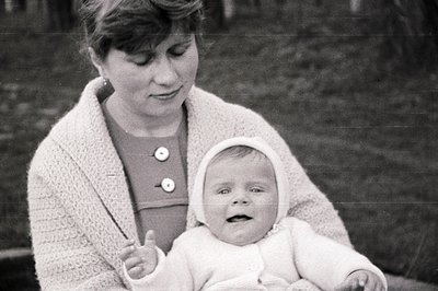 A mother and baby in a tender, intimate portrait. The mother wears a textured sweater and button-down dress, cradling an infa...