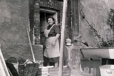 A woman stands in a weathered stone doorway, apron adorned with a patterned print. A woven basket, wooden crates, and a metal...