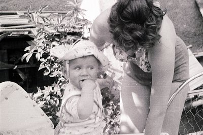 A young child with a patterned sun hat sits outdoors, appearing to eat something, while a woman leans in closely. A glimpse o...