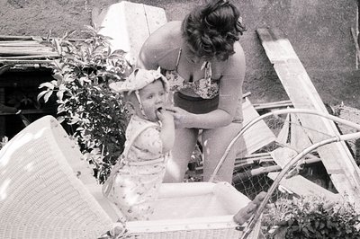 A young child sits in a wicker chair, appearing to receive a treat from a woman in a floral bathing suit. The scene is set ou...