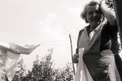 A woman in a dark dress and patterned apron hangs laundry on a line outdoors. She holds a sheet, appearing to shield her eyes...