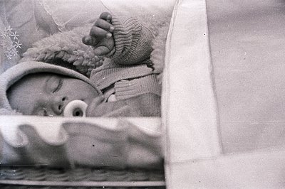 Infant sleeps peacefully bundled in a knitted bonnet and textured clothing. Vintage photograph likely from the mid-20th centu...