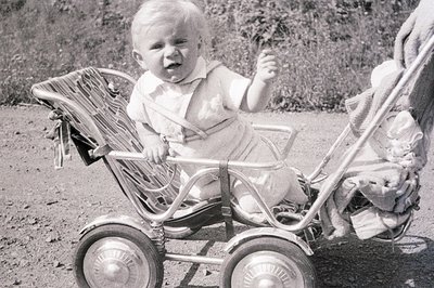 An infant sits in a vintage, wire-spoke stroller, raising one hand. The child wears a light-colored, collared outfit. Backgro...