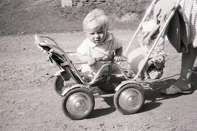 A young child sits in a vintage, chrome-framed tricycle with balloon tires. The child wears a short-sleeved shirt and suspend...