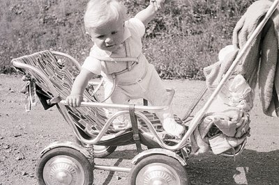 A young child, appearing to be a toddler, is playfully posed on a vintage stroller outdoors. The child is clad in a light-col...