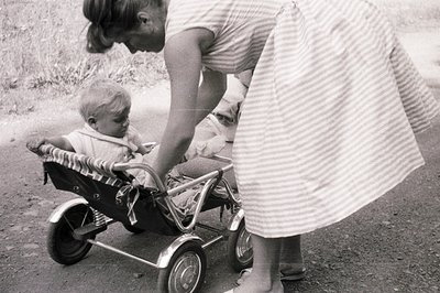A young boy sits in a doll carriage, being gently steered by a woman in a striped dress. The scene, likely a backyard or driv...