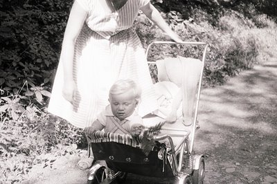 A young boy sits in a vintage, chrome-framed stroller, appearing slightly distressed. A woman in a patterned dress guides the...