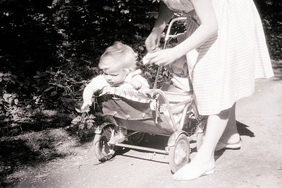 A black and white photo shows a young child seated in a vintage, metal baby carriage on a paved path. The child, wearing a sh...
