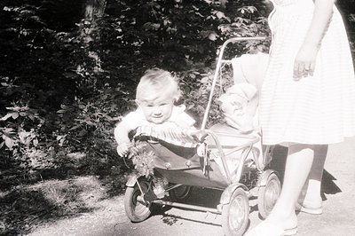 A young child sits in a vintage metal stroller, appearing to cry or express a strong emotion. A baby in a bonnet rests within...