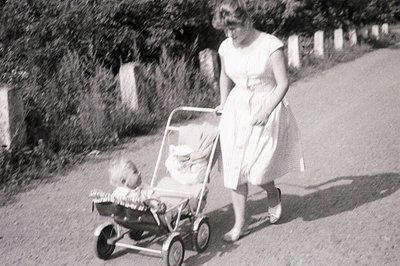 A woman in a light-colored dress pushes a baby in a vintage pram along a gravel road, framed by a stone wall and lush foliage...