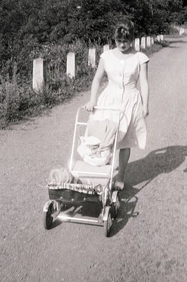 A young woman in a light-colored, patterned dress pushes a vintage silver baby carriage along a gravel road. The carriage is ...