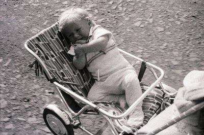 A young child rests in a wicker-framed stroller with padded seat, wearing a short-sleeved romper. The image, likely from the ...