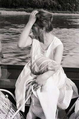 A woman in a striped dress sits outdoors, seemingly on a boat, with a blurred lakeside view. She appears to be pushing her ha...