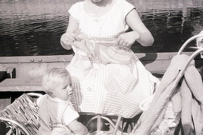 A young girl in a striped dress sits in a wicker stroller, focused on a cloth doll or toy. A toddler sits in the stroller's l...