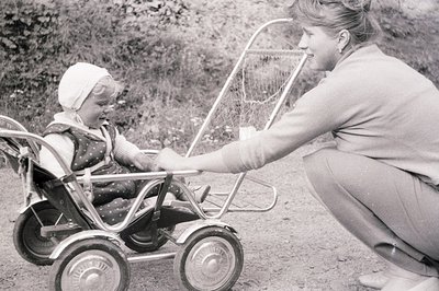 A woman kneels beside a young child seated in a vintage baby carriage with a wire basket. Child wears a bonnet and patterned ...