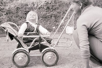 A young child sits serenely in a vintage, chrome-framed stroller. A woman kneels nearby, offering a gentle interaction. The s...