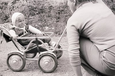 A toddler sits in a vintage, metal doll stroller, pulled by a caregiver in a long-sleeved sweater. The scene depicts a domest...