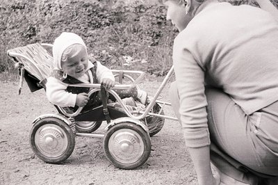 A toddler in a vintage metal stroller, wearing a bonnet and overalls, grins. An adult, possibly a caregiver, is crouched near...
