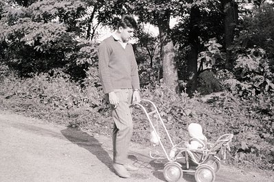A young man in a sweater and trousers strolls along a gravel road, pushing a vintage doll carriage. Likely mid-20th century, ...