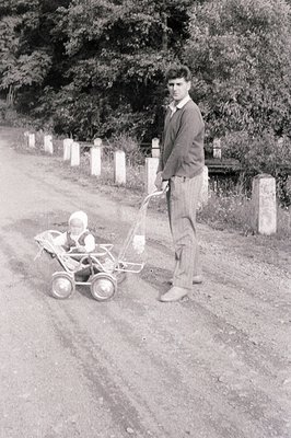 A young man in a tweed sweater and trousers stands beside a vintage stroller with a baby bundled within. The gravel road and ...